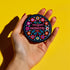 Round black pinback button with vibrant floral embroidery design surrounding bold pink and red text reading "Dissent is Patriotic."